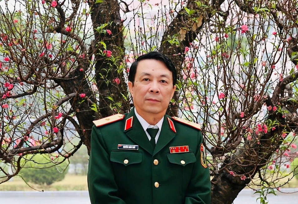 Screenshot20260419181636996com android chrome catrawarta Portrait of a man in a green military uniform standing outdoors in front of a pink flowered blossoming tree